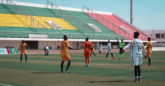 Jogadores de futebol em campo durante uma partida no Girabola, com bancadas coloridas ao fundo.