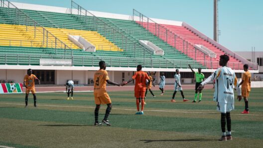 Jogadores de futebol em campo durante uma partida no Girabola, com bancadas coloridas ao fundo.