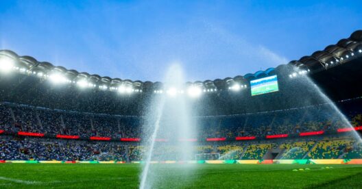 Vista noturna de um estádio de futebol com irrigadores irrigando o campo, realçando a atmosfera vibrante.