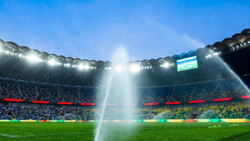 Vista noturna de um estádio de futebol com irrigadores irrigando o campo, realçando a atmosfera vibrante.