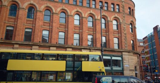 Autocarro amarelo 'BEE NETWORK' e táxi preto em frente a um edifício de tijolo em Manchester.