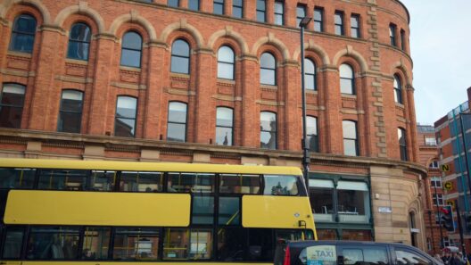 Autocarro amarelo 'BEE NETWORK' e táxi preto em frente a um edifício de tijolo em Manchester.
